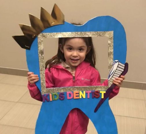 Little girl holding a sign shaped like a tooth reading Kids Dentist